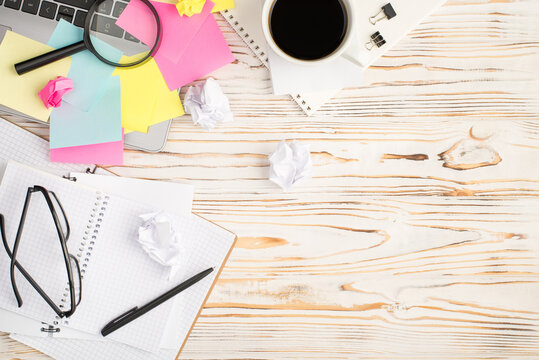 Top view photo of cup of coffee binder clips multicolor sticker note paper magnifier laptop glasses pen notebooks and crumpled paper balls on isolated bright wooden table background with copyspace