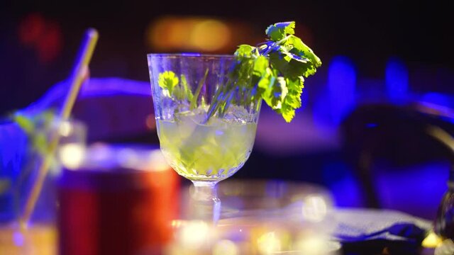parallax panning shot shoiwng a mocktail drink placed on a table with mint, lime, stem glass with a person talking behind and colorful lights and out of focus backgrounds in a cafe pub club night life