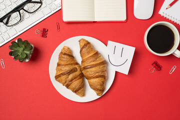 Top view photo of workplace keyboard mouse stationery glasses flowerpot copybooks cup of coffee plate with two fresh croissants and sticky note paper with drawn smiling face on isolated red background