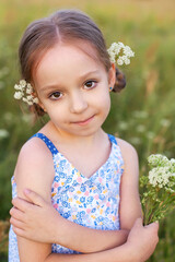 A little blonde girl walks through a summer wheat field in a long blue sundress. The child smiles and looks at the camera.