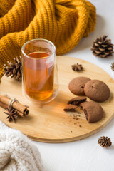 Vertical photo of winter composition glass cup of hot tea bitten off chocolate cookies anise and cinnamon sticks on wooden tray pine cones orange sweater and white scarf