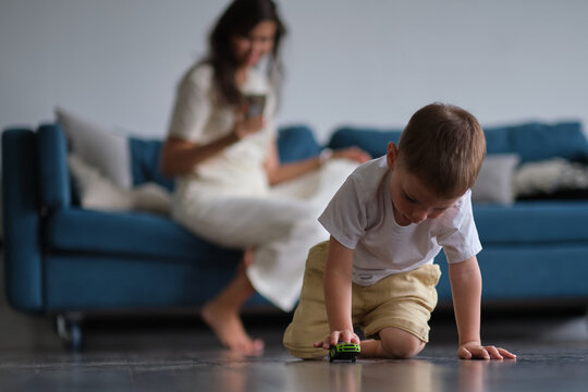 Image of young pretty little boy playing on the floor with car toys and drawing while his mother sitting on a sofa.