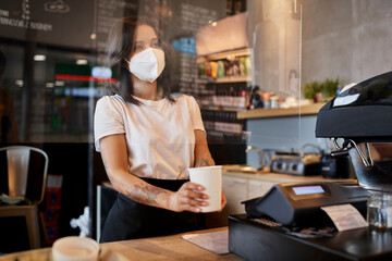 Fototapeta na wymiar Female barista at cafe giving coffee cup through protective plastic screen wearing mask