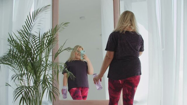 Blonde confident overweight woman walks to a mirror and taking a picture of herself on the phone