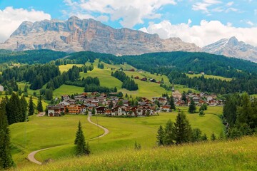 Fototapeta na wymiar Summer scenery of a beautiful village at foothills of rugged Sasso di Santa Croce & a country road winding in the green grassy valley in Val Badia, Alta Badia, Dolomiti, Trentino, South Tyrol, Italy