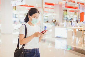 Asian woman wearing face mask using a smartphone in shopping mall. Masked woman in white t-shirt with face mask in mall . Coronavirus or covid-19 concept. Bokeh background.