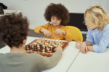 Awesome little children discussing game while sitting at the table and playing chess indoors