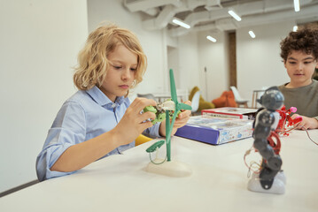 Create things. Caucasian boy examining technical toy while sitting at the table together with other...