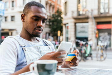 african young man looking to his credit card and his smart phone. He is sitting on a chair in a bar terrace