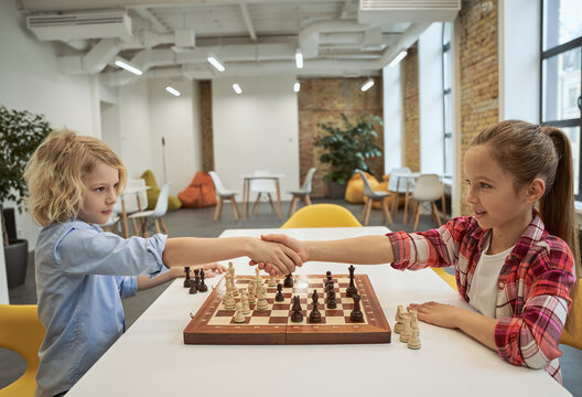 Respect. Adorable little friends, caucasian boy and girl shaking hands after match, playing chess, sitting together at the table in school
