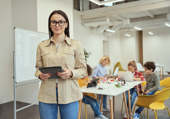Beautiful young female teacher in glasses looking at camera and holding tablet pc while standing in a classroom. Kids working on diy robot in the background