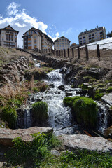 Water from a stream that falls from above with grass and rocks, next to a fence with mountain houses in the background