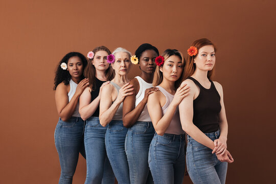 Six diverse female standing together over brown background. Women of different ages with flowers in their hair.