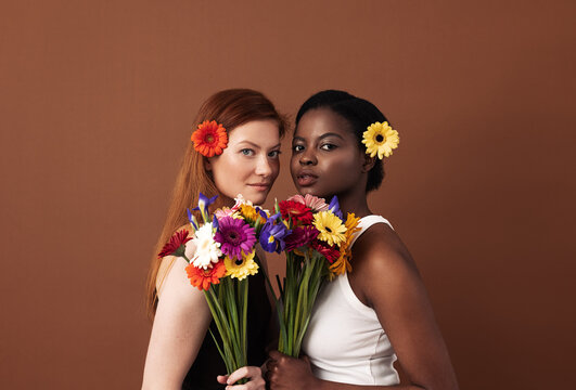 Two women of different races with flowers in their hairs holding bouquets. Caucasian and African American females looking at camera in a studio.