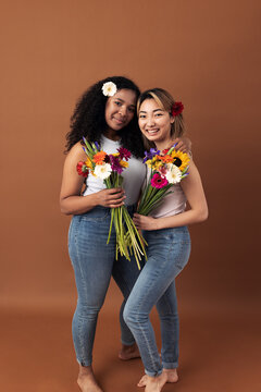 Asian and mixed race women posing against a brown background. Two smiling females with bouquets looking at camera.