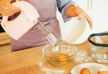 Young woman baking a cake in the kitchen, using a mixer to whisk the fresh ingredients in a glass mixing bowl.