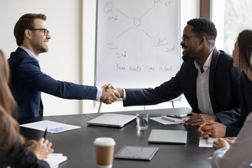 Happy multiethnic diverse business leaders shaking hands over meeting table in training room. Boss congratulating employee on hiring, promotion, work achieve, expressing recognition, gratitude
