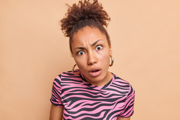 Close up shot of shocked concerned indignant curly woman wears earrings striped t shirt hears something unexpected poses against brown background. Human face expressions and reactions concept