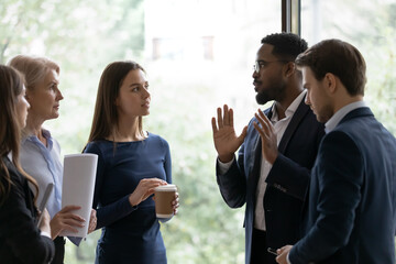 Diverse business team standing, talking at office window, brainstorming on project at casual meeting. African American group leader speaking to employees on briefing. Interns discussing training exam