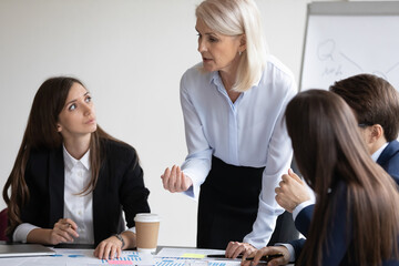 Confident senior business teacher training group of interns, explaining work on marketing charts to employees on training meeting. Serious businesswoman talking to team, discussing project reports