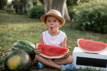 Kid with hat smiling very emotional and eating watermelon. Funny child hold fruit. Outdoors in park, summers season of fruits and berries. healthy food