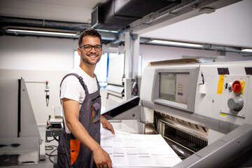 Portrait of smiling caucasian operator standing by paper cutting machine in printing factory. - obrazy, fototapety, plakaty