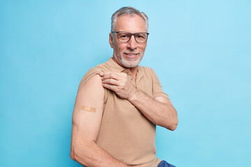 Old man with grey hair shows vaccinated arm motivates to vaccinate against coronavirus to stop epidemic cares about health in his age wears brown t shirt spectacles isolated over blue background - obrazy, fototapety, plakaty