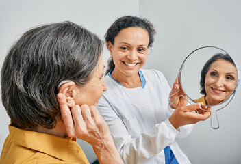 Mature woman with BTE hearing aid looks at himself in mirror held by smiling audiologist, and tries...