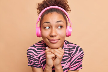 Close up portrait of thoughtful pleased dark skinned woman keeps hands under chin wears headphones on ears listens audio track or tutorial lecture concentrated aside isolated over beige wall