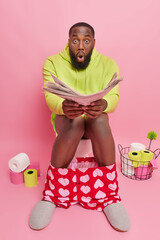 Vertical shot of shocked bearded Afro American man with boxers down on legs reads newspaper to find...