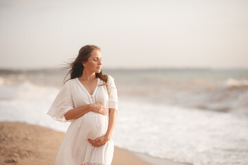 Smiling beautiful pregnant woman wear stylish white dress hold belly walk at beach over waves of...