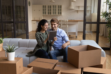 Planning renovations. Happy young wife sit on sofa at new house show to husband interior design studio website on tablet pc. Loving couple sit among boxes at moving day look at home decor ideas at web