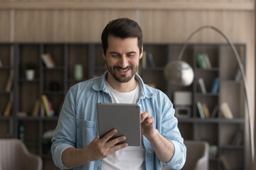 Happy young caucasian man holding digital computer tablet in hands, enjoying web surfing information, communicating distantly in social network, playing online games or using software applications.