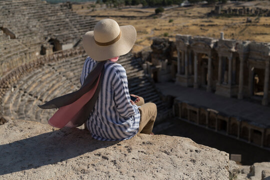 Tourist wearing hat sitting in ancient theater of Hierapolis taking photo.