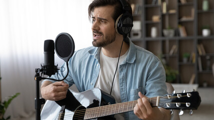 Happy young male musician in headphones singing song in stand microphone, playing guitar, creating recording own music, practicing indoors using professional equipment, hobby activity concept.