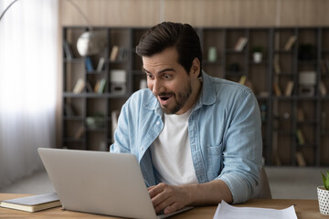 Surprised young male manager worker looking at laptop screen, feeling excited of getting email with amazing news, getting dream job offer or promotion notification, celebrating salary increase.