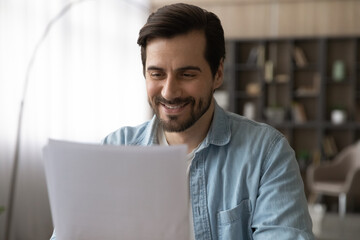 Happy millennial generation man reading paper document, feeling excited of getting good news notification, bank loan approval or paid taxes refund notice. Male accountant reviewing financial report. - obrazy, fototapety, plakaty