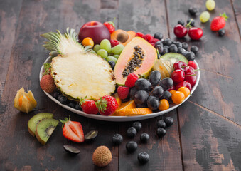 Fresh raw organic summer berries and exotic fruits in white plate on dark wooden background. Pineapple, papaya, grapes, nectarine, orange, apricot, kiwi, strawberry, lychees, cherry and physalis.