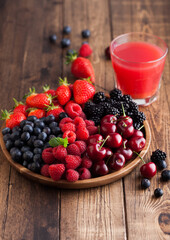 Fresh organic summer berries mix in round wooden tray with glass of juice on light wooden table background. Raspberries, strawberries, blueberries, blackberries and cherries.