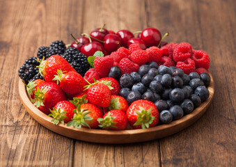 Fresh organic summer berries mix in round wooden tray on light wooden table background. Raspberries, strawberries, blueberries, blackberries and cherries.
