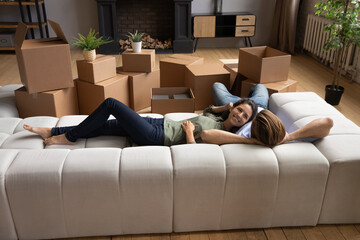 Smiling wife lie on couch cuddle with beloved husband rest after moving relocation to new modern house. Happy young couple in love take break in remodeling renovating home rest close to boxes packages