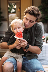 Small boy eat watermelon sit on father knees outside house in garden or near camper car while road...