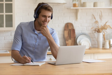 Confident smiling young male student in modern headset watch training web course from home take...
