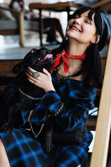 Happy woman hold adorable french bulldog on lap in cafeteria. Cheerful trendy female enjoy spend time with pet in cafe. Young joyful girl in fashion clothes hug black cute dog and laugh with happiness