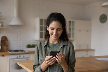 Communication on distance. Happy young hispanic lady stand at modern kitchen interior look at phone...