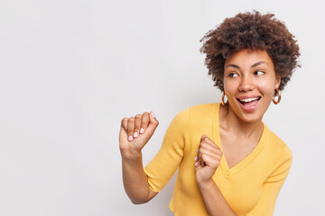 Positive carefree young woman shakes fists moves with rhythm of music feels happy dances and looks away wears casual yellow jumper isolated over white background copy space for your information