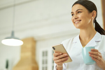 Happy female doctor standing with smartphone and cup