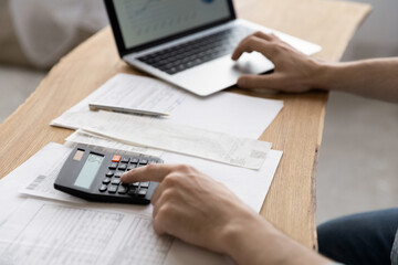 Close up young man using calculator summarizing domestic utility bills or taxes, managing household monthly budget, planning investment, reviewing financial payments alone at home, using computer app.
