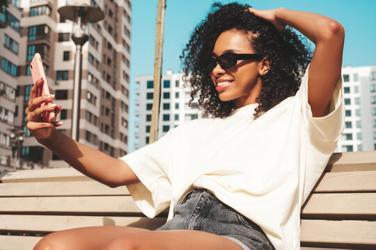 Beautiful black woman with afro curls hairstyle.Smiling hipster model in white t-shirt. Sexy carefree female posing in the street in sunglasses. Cheerful and happy.Taking selfie photo