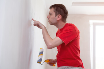 Man with roller touch and check up the wall after work. Repair of the interior. Young male decorator painting or primer a wall.